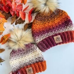 Two Chunky Suzette Stitch Beanies with faux fur pom-poms in autumn colors, placed next to red-orange leaves on a white surface.