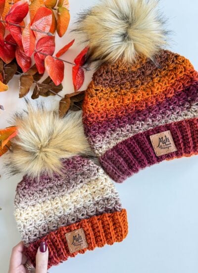 Two Chunky Suzette Stitch Beanies with faux fur pom-poms in autumn colors, placed next to red-orange leaves on a white surface.