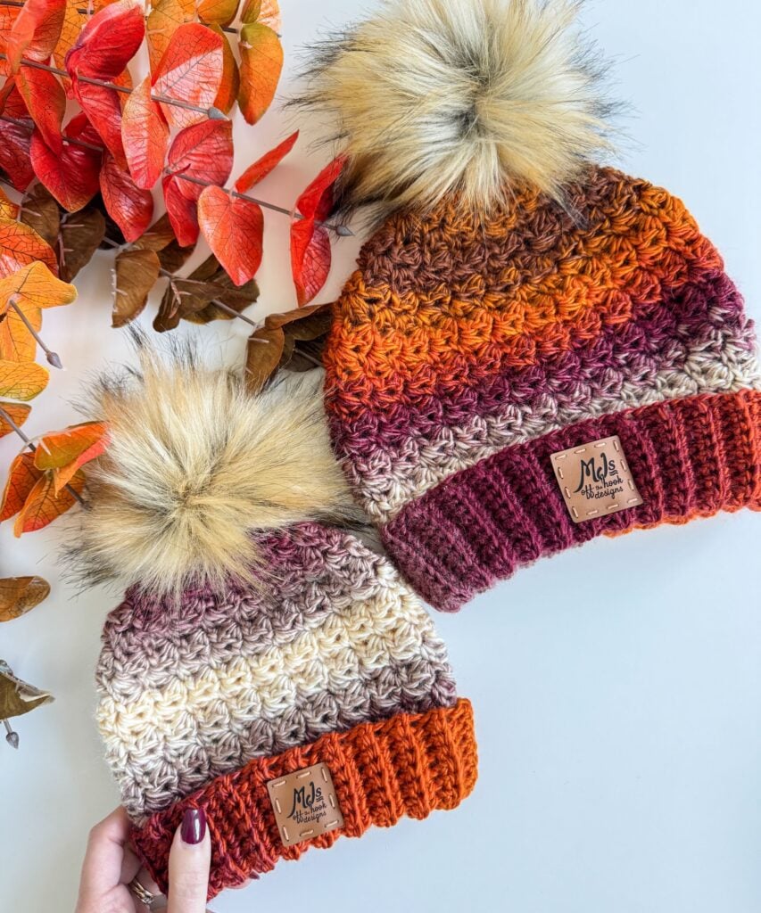 Two Chunky Suzette Stitch Beanies with faux fur pom-poms in autumn colors, placed next to red-orange leaves on a white surface.