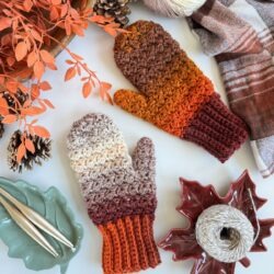 A pair of crocheted fingerless gloves and mittens in autumn colors is displayed on a white surface, surrounded by yarn, crochet hooks, pinecones, leaves, and a plaid scarf.