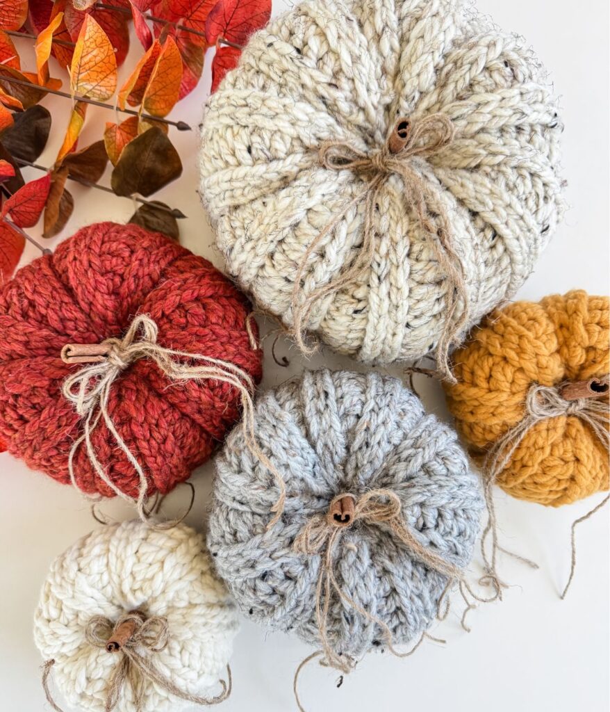 Five Harvest Pumpkin-inspired knitted pumpkins in white, red, gray, cream, and mustard yellow are arranged on a white surface next to orange autumn leaves.