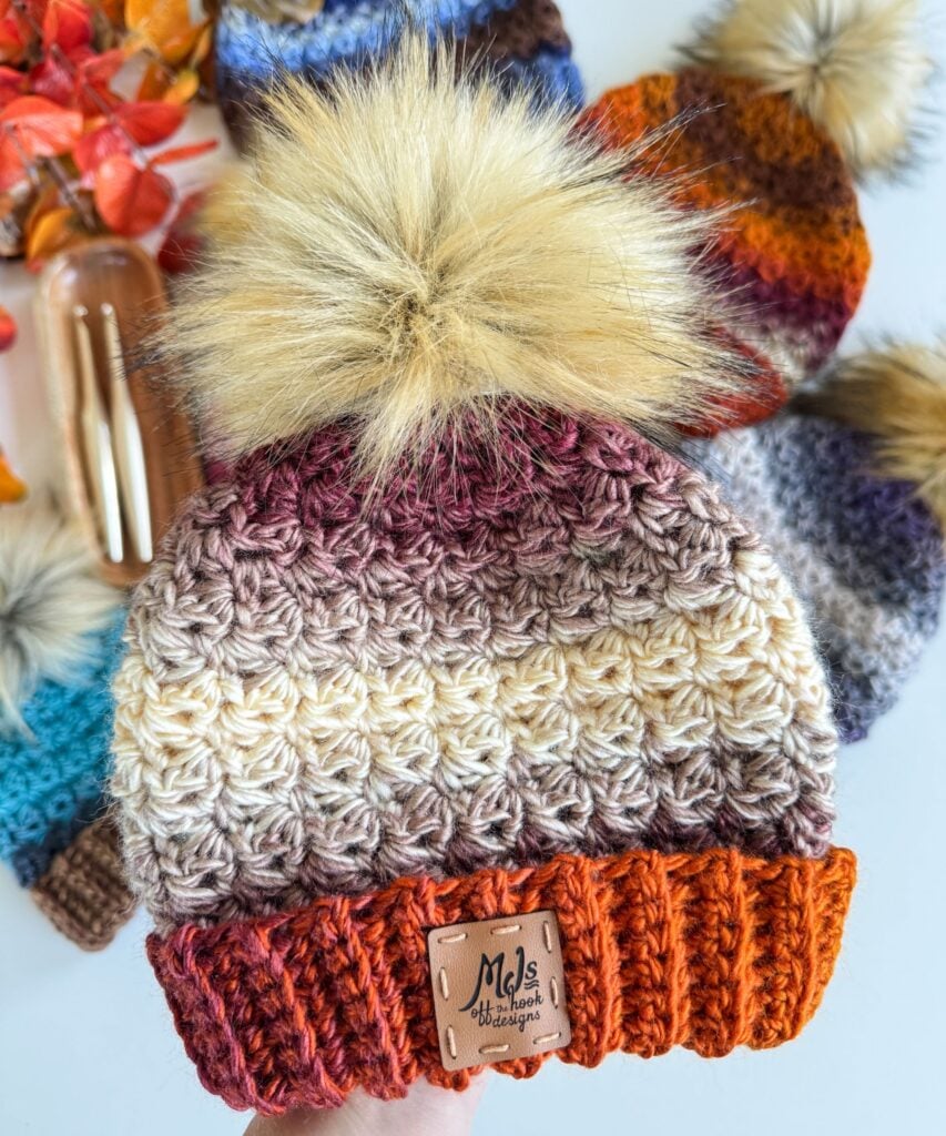 Hand holding a crocheted, multicolored Chunky Suzette Stitch Beanie with a large faux fur pom-pom; other similar hats and crochet hooks are visible in the background.