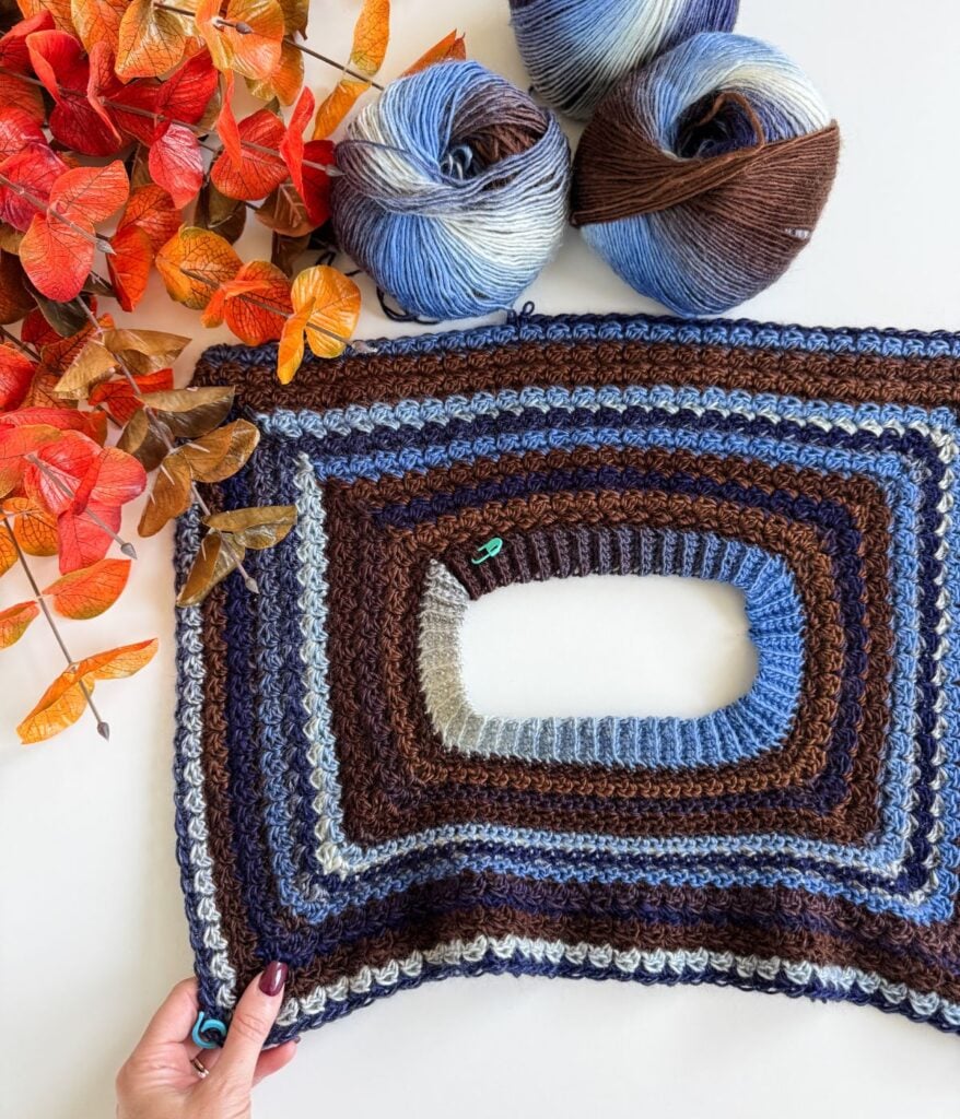 Rectangular crochet project in shades of blue and brown, crafted with Suzette Stitch Raglan technique, featuring an oval opening in the center, two balls of yarn, and orange artificial leaves on a white surface.