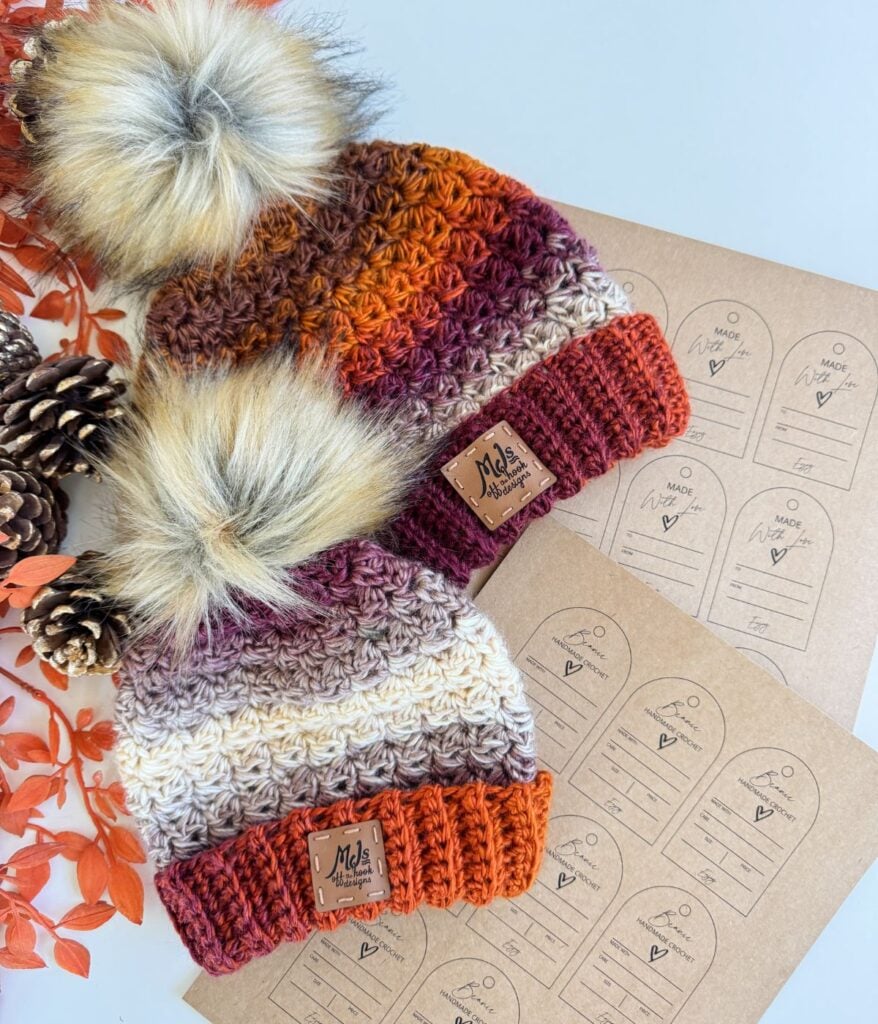 Two Chunky Suzette Stitch Beanies with faux fur pom-poms are placed beside pinecones, orange leaves, and kraft paper product tags on a white surface.