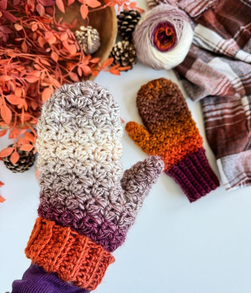 A hand wearing a crocheted autumn-colored mitten holds another mitten; nearby, fingerless gloves, yarn, pinecones, dried leaves, and a plaid cloth complete the cozy scene.