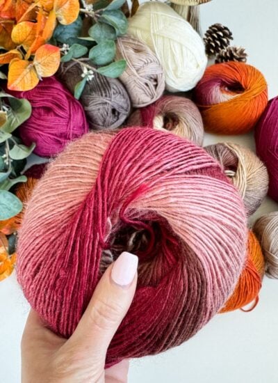 A hand with light pink nail polish holds yarn for a Chunky Suzette Stitch Beanie, hovering above other colorful yarn balls, with autumn leaves and pinecones nearby.