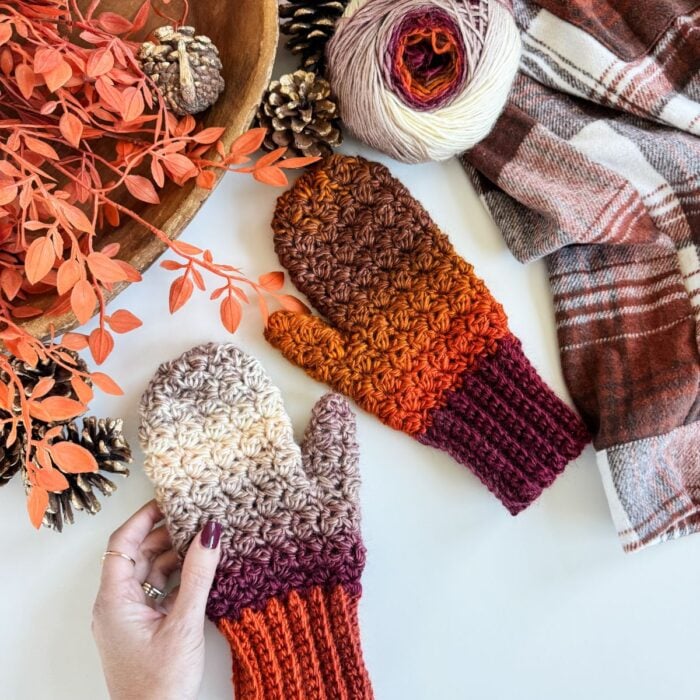 A hand holds a crocheted mitten with ombre shades from cream to burgundy, next to another mitten, fingerless gloves, yarn, pinecones, orange foliage, and a plaid scarf on a white surface.