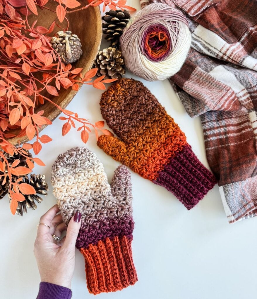 A hand holds a crocheted mitten with ombre shades from cream to burgundy, next to another mitten, fingerless gloves, yarn, pinecones, orange foliage, and a plaid scarf on a white surface.