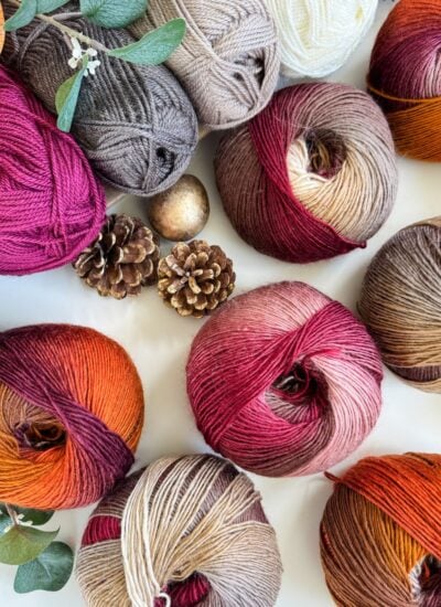 Balls of multicolored yarn, pinecones, leaves, and decorative spheres are arranged on a white surface, evoking cozy autumn vibes perfect for crafting a Chunky Suzette Stitch Beanie.