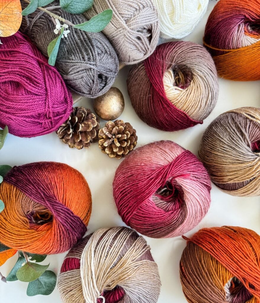 Balls of multicolored yarn, pinecones, leaves, and decorative spheres are arranged on a white surface, evoking cozy autumn vibes perfect for crafting a Chunky Suzette Stitch Beanie.