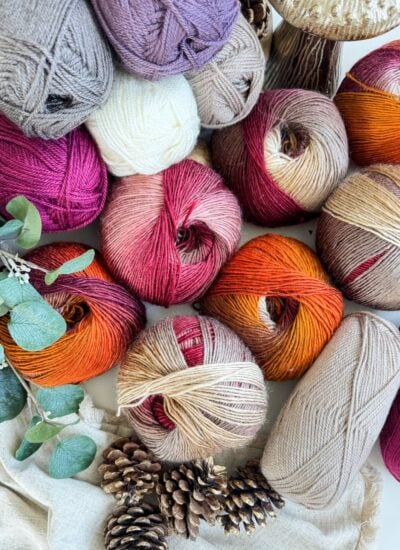 Assorted yarn balls in shades of red, orange, tan, and cream are arranged with pine cones, greenery, and a neutral cloth—perfect for crafting cozy fingerless gloves and mittens.
