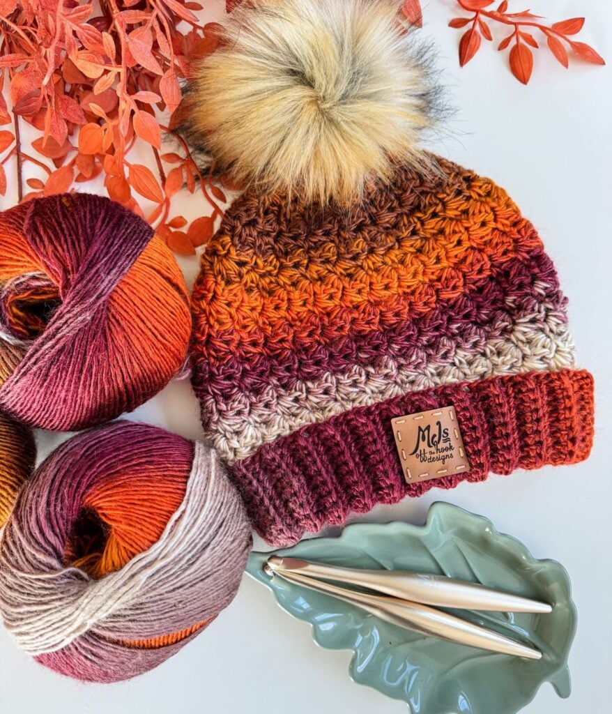 Crocheted Chunky Suzette Stitch Beanie with a faux fur pompom in autumn colors, two matching yarn balls, metal crochet hooks, and a leaf-shaped dish on a white background scattered with red leaves.
