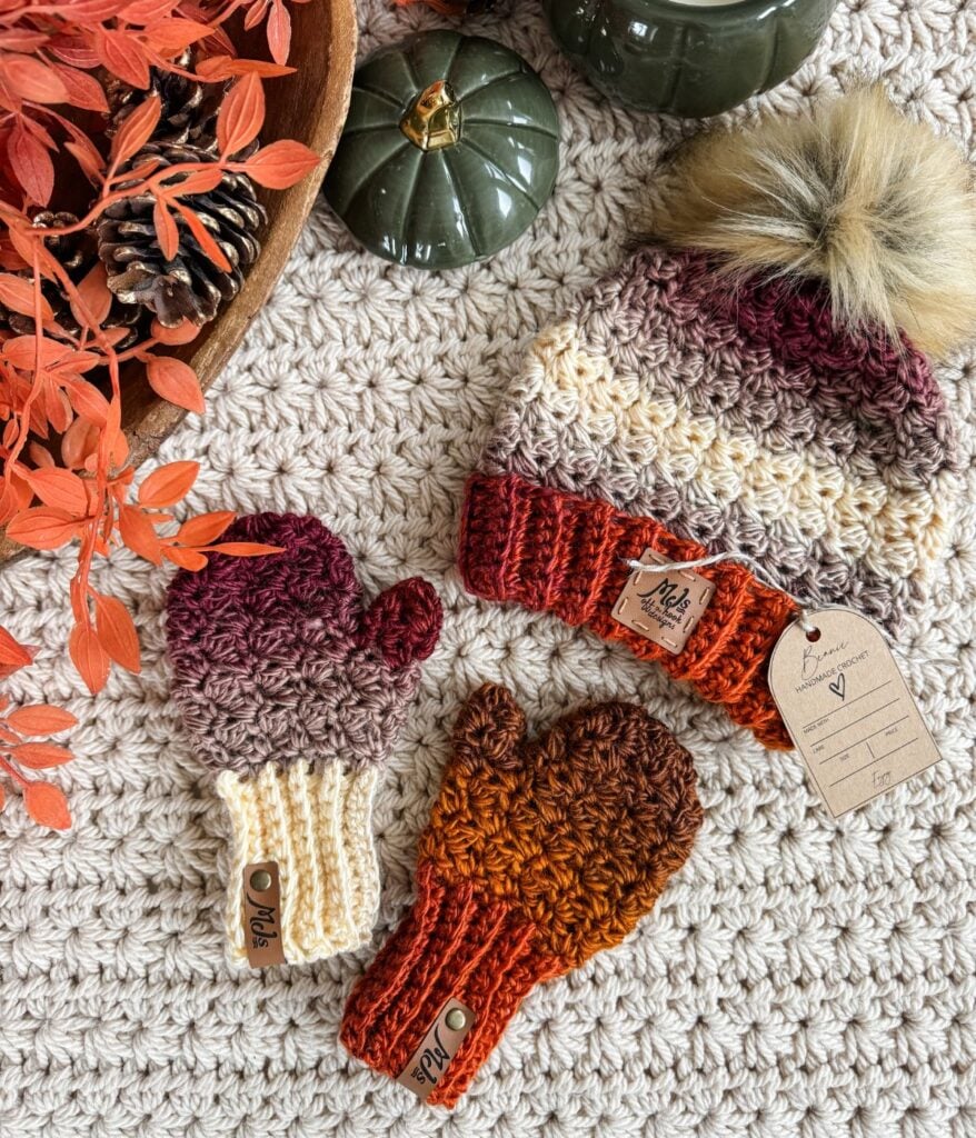 A knitted hat with a faux fur pom-pom, two pairs of fingerless gloves and mittens are displayed on a textured beige surface, alongside autumn leaves, a pinecone, and green ceramic pumpkins.