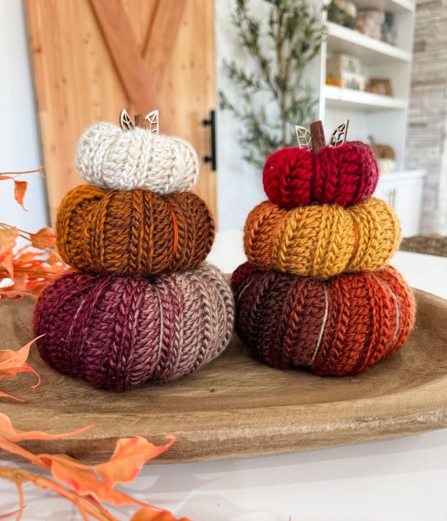 Two stacks of Harvest Pumpkin knitted pumpkins in fall colors sit on a wooden tray, with autumn leaves and home decor visible in the background.
