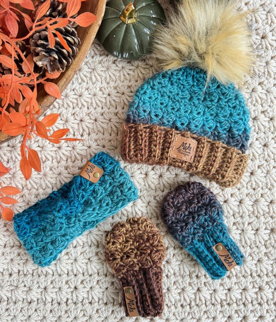 A crocheted blue and brown hat with a faux fur pom-pom, matching ear warmer, and two keychain mittens, plus cozy fingerless gloves—all with branded tags—displayed on a textured surface with autumn decor.