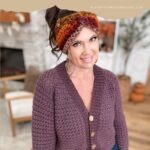Woman wearing a textured purple Suzette Raglan crochet cardigan and a multicolored knit headband stands indoors near a fireplace and wooden chair. Text overlay promotes the Suzette Raglan crochet pattern and video.