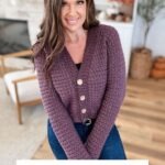 Woman wearing a textured purple Suzette Raglan cardigan with gold buttons stands indoors, smiling. Text reads: "The Suzette Raglan Cardigan Crochet Pattern & Video.