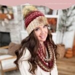 Woman wearing a Chunky Suzette Stitch Beanie with a pom-pom and a matching cowl scarf, standing indoors in a cozy, decorated living room.