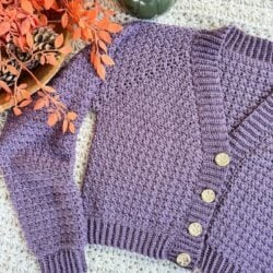 A purple crocheted Suzette Raglan cardigan with four gold buttons is laid on a textured surface near an arrangement of orange leaves and pinecones.