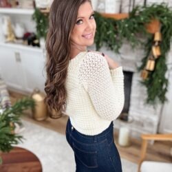 A woman with long brown hair in a cream Suzette Stitch Raglan sweater and dark jeans stands indoors, smiling over her shoulder in a room decorated with greenery.