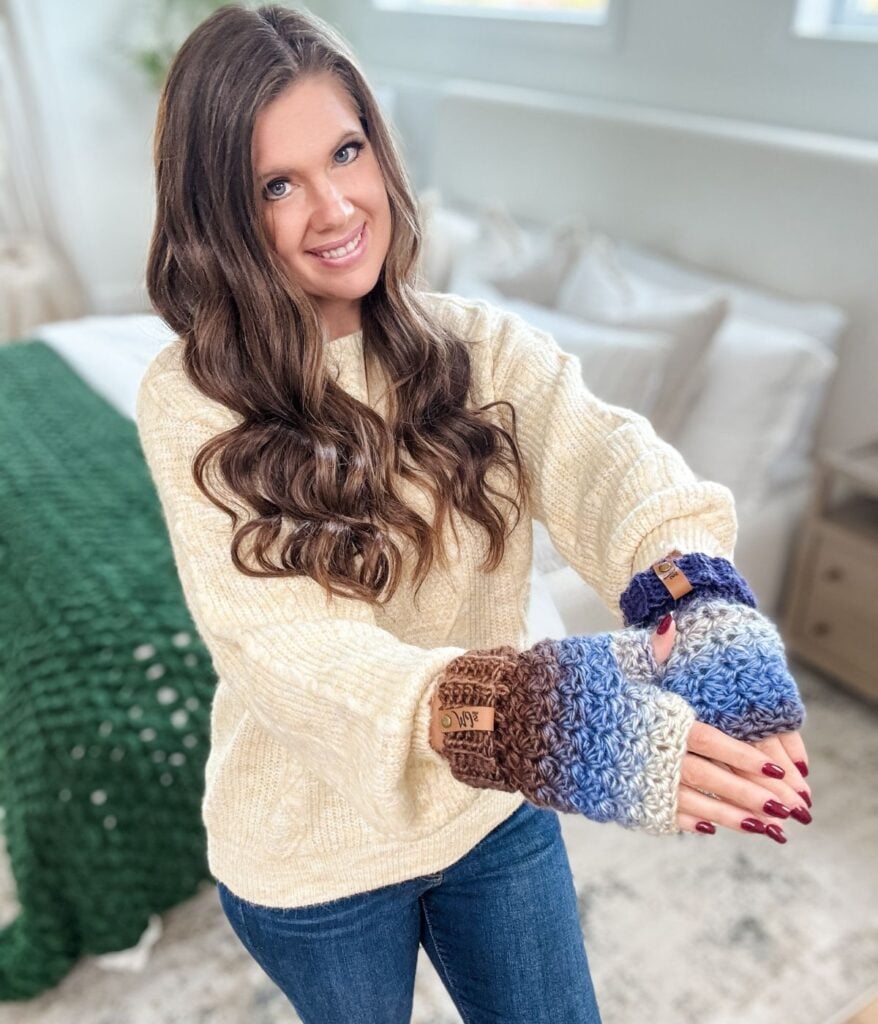 Woman with long brown hair wearing a cream sweater and blue jeans, showing off multicolored crocheted fingerless gloves and mittens in a cozy bedroom setting.