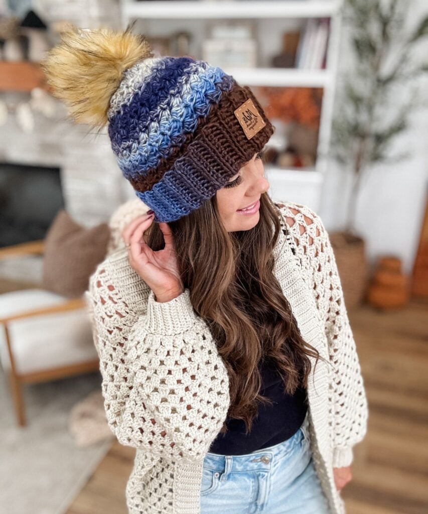 A woman wearing a Chunky Suzette Stitch Beanie with a pom-pom and a cream-colored crochet cardigan stands indoors, looking to the side.