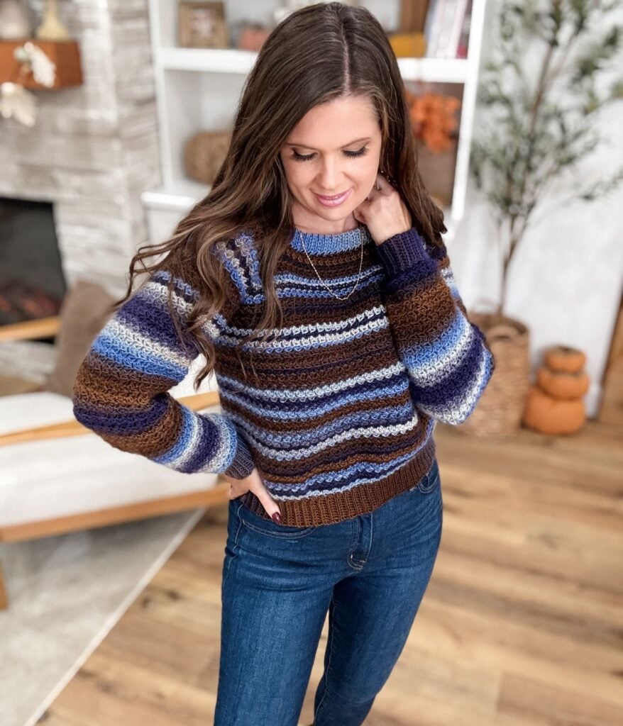 Woman standing indoors wearing a Suzette Stitch Raglan, a striped long-sleeve sweater in shades of blue and brown with blue jeans, looking down with one hand on her hip.