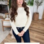 Woman wearing a cream-colored Suzette Raglan crochet sweater stands indoors, smiling at the camera. Text below reads: "Suzette Stitch Raglan Sweater Crochet Pattern & Video.
