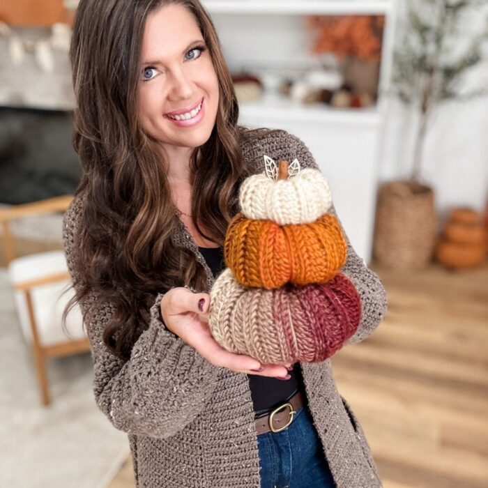 Woman holding a stack of three Harvest Pumpkin-inspired knitted pumpkins in cream, orange, and red, standing in a cozy, autumn-themed living room.