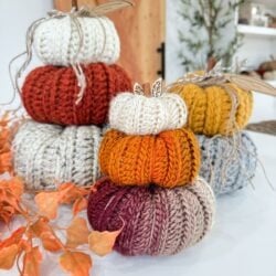 Three stacks of Harvest Pumpkin knitted pumpkins in various autumn colors are displayed on a white surface, with orange leaves and plants in the background.