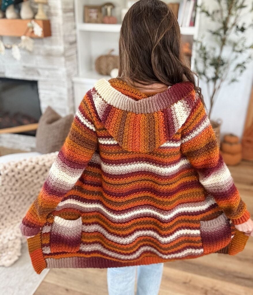 A woman with long brown hair is seen from the back inside a cozy room, wearing a striped, multicolored crocheted Suzette Raglan cardigan with pockets.