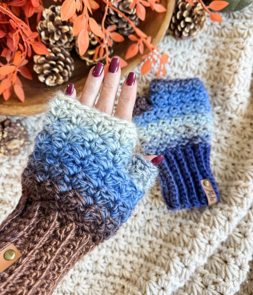 A hand wearing a blue and brown crocheted fingerless glove rests on a textured cream blanket, with matching fingerless gloves and mittens and autumn decorations nearby.