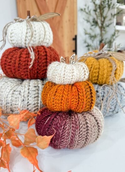 Three stacks of Harvest Pumpkin knitted pumpkins in various autumn colors are displayed on a white surface, with orange leaves and plants in the background.