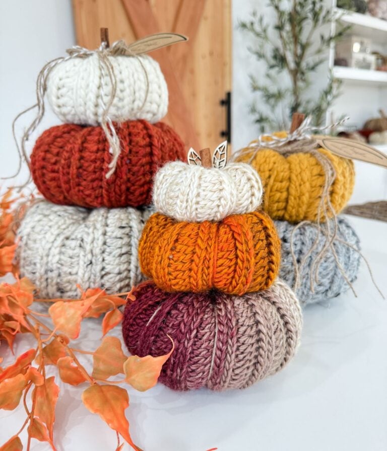 Three stacks of Harvest Pumpkin knitted pumpkins in various autumn colors are displayed on a white surface, with orange leaves and plants in the background.