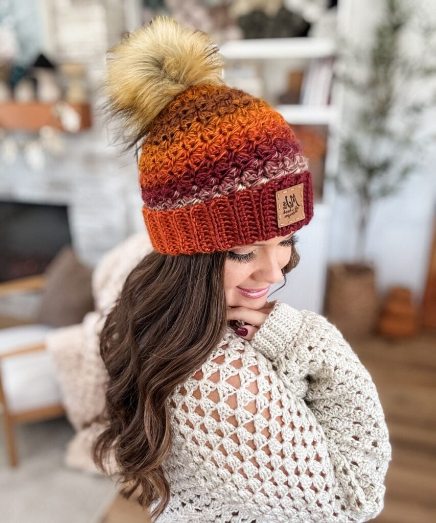 Woman in a crochet sweater wearing a Chunky Suzette Stitch Beanie with a faux fur pom-pom, standing indoors and smiling with her head turned to the side.