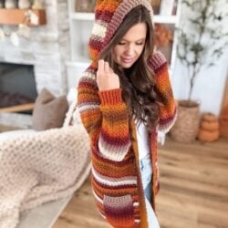 Woman indoors wearing a Suzette Raglan striped cardigan in autumn colors, with light denim jeans, standing on a wooden floor near a fireplace and shelves.