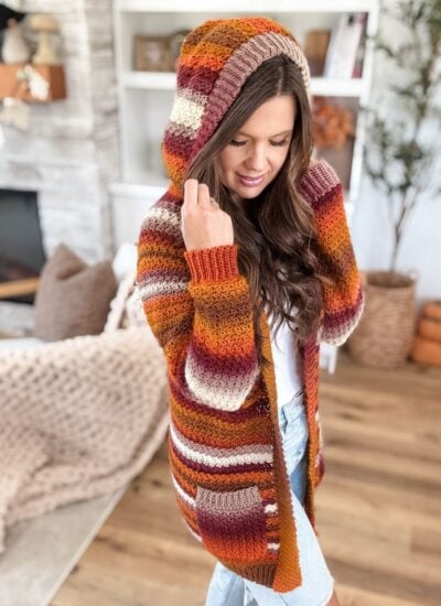 Woman indoors wearing a Suzette Raglan striped cardigan in autumn colors, with light denim jeans, standing on a wooden floor near a fireplace and shelves.