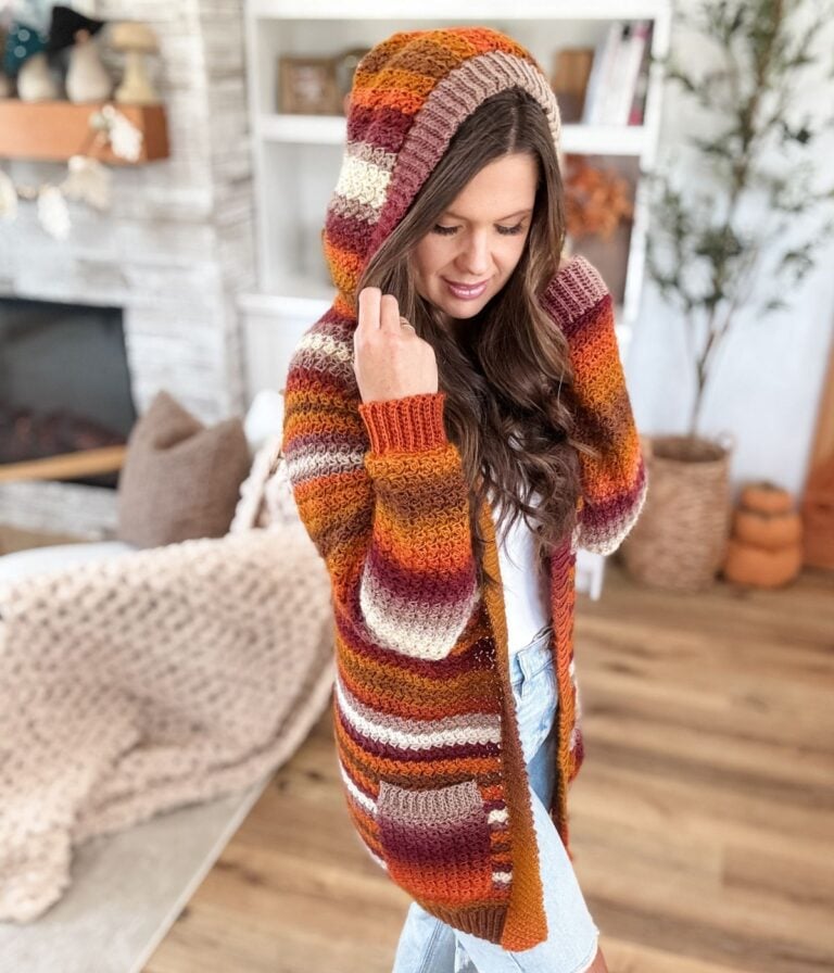 Woman indoors wearing a Suzette Raglan striped cardigan in autumn colors, with light denim jeans, standing on a wooden floor near a fireplace and shelves.