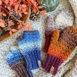 Two pairs of crocheted fingerless gloves and mittens—one fingerless in blue and brown, one full in orange and brown—are arranged on a textured surface with cozy autumn decor.