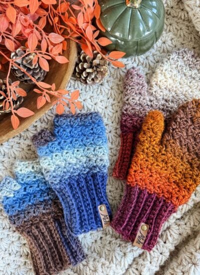 Two pairs of crocheted fingerless gloves and mittens—one fingerless in blue and brown, one full in orange and brown—are arranged on a textured surface with cozy autumn decor.