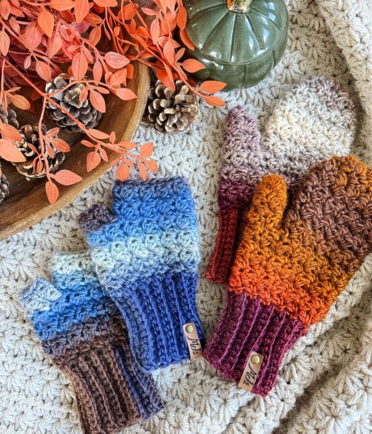 Two pairs of crocheted fingerless gloves and mittens—one fingerless in blue and brown, one full in orange and brown—are arranged on a textured surface with cozy autumn decor.