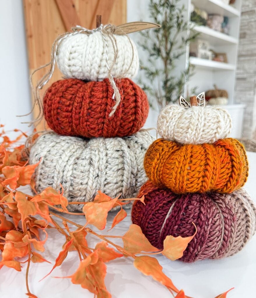 Two stacks of knitted harvest pumpkin decor in fall colors are displayed on a table, with orange leaves in the foreground and shelves in the background.