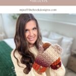 A woman with long brown hair wearing a cream sweater and colorful crochet fingerless gloves and mittens sits on a bed, smiling at the camera.