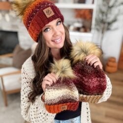 A woman wearing a Chunky Suzette Stitch Beanie with a faux fur pom-pom holds two similar beanies in her hands while standing in a cozy, decorated living room.