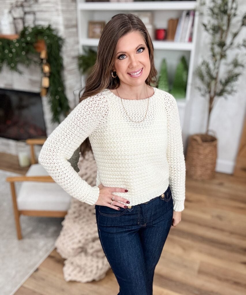 A woman with long brown hair wearing a cream Suzette Stitch Raglan sweater and dark jeans stands indoors, smiling at the camera. The background includes a fireplace, shelves, and plants.