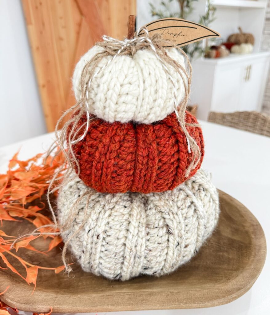 A stack of three Harvest Pumpkin knitted pumpkins in cream, orange, and beige sits on a wooden tray, with an autumn leaf decoration beside it.