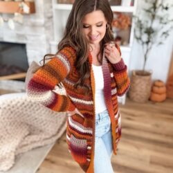 A woman wearing a striped, autumn-colored Suzette Raglan cardigan over a white top and light blue jeans stands indoors on a wooden floor, smiling and looking down.