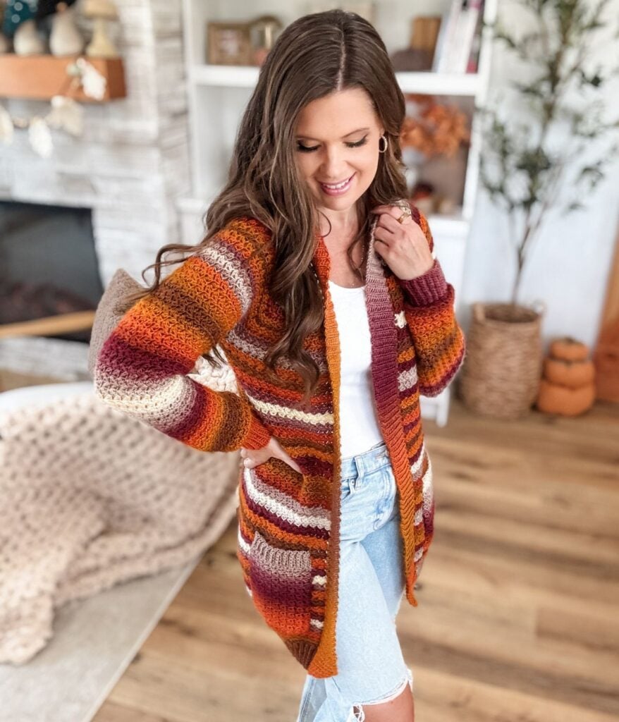 A woman wearing a striped, autumn-colored Suzette Raglan cardigan over a white top and light blue jeans stands indoors on a wooden floor, smiling and looking down.