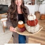 A woman holding a tray of harvest pumpkins in various colors stands indoors. Text on the image reads: "Chunky Harvest Pumpkins Crochet Pattern & Video.