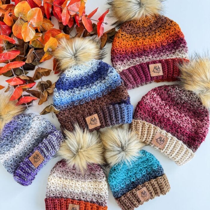 Five Chunky Suzette Stitch Beanie hats with pom-poms, each in different multicolored stripes, are arranged next to orange and red artificial leaves on a white background.